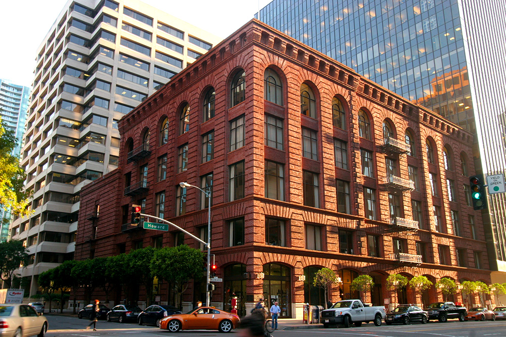A historic red brick building on a city street corner, surrounded by modern office buildings