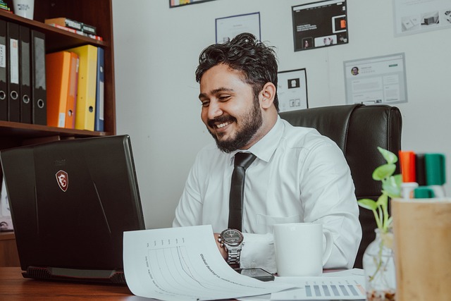Smiling businessman in a white shirt and black tie working at a desk with a laptop, printed charts, a coffee mug, and office supplies, appearing engaged and professional in a modern office setting.
